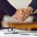 Mãos de duas pessoas apertando em um acordo, com óculos e notebook ao fundo.